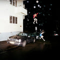 Two people in costumes leaping in the air over a car on a dark street.