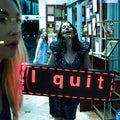 A woman in a sparkling black dress holds a large LED sign that reads 'I quit' in red light inside what appears to be a shop or office. Another person is partially visible in the foreground, and another person stands in the background near framed pictures and shelves.