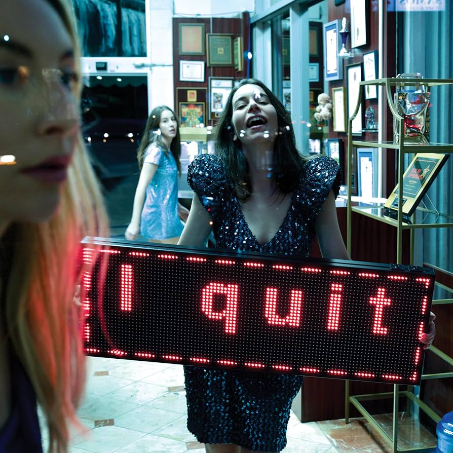 A woman in a sparkling black dress holds a large LED sign that reads 'I quit' in red light inside what appears to be a shop or office. Another person is partially visible in the foreground, and another person stands in the background near framed pictures and shelves.