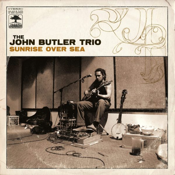 Album cover for Sunrise Over Sea by John Butler Trio featuring a warm-toned coastal photograph with the band standing on a beach at dawn, capturing a relaxed, organic atmosphere.