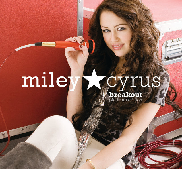 Album cover for Breakout Platinum Edition by Miley Cyrus showing Miley seated against red road cases, holding a red microphone, with long wavy brown hair and a confident smile.