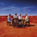 Four people sitting around a table in a desert landscape with red sand and blue sky.