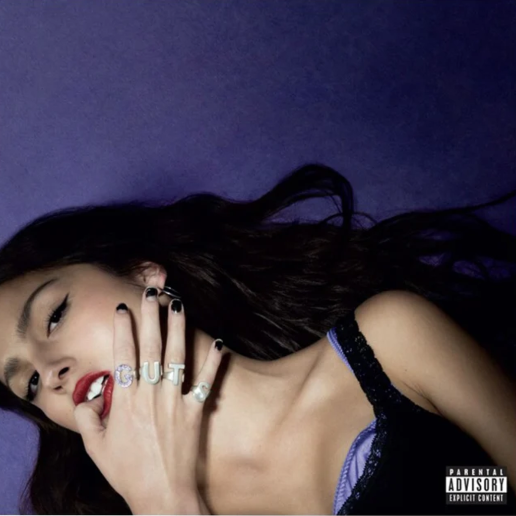 Cover art for Guts by Olivia Rodrigo featuring a photo of Olivia laying on a purple background with rings on her fingers spelling out the word 'Guts'.