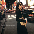 Woman standing on a city street at night with neon lights and cars in the background, holding a beige bag.