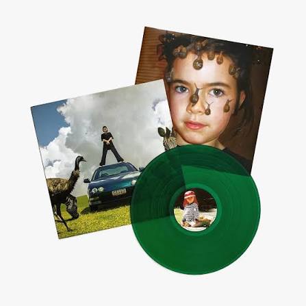 A green translucent vinyl record partially covering a square album cover, with another album cover behind it showing a child's face with brown spots and a person standing on a car outdoors. The product appears to be a music album package.